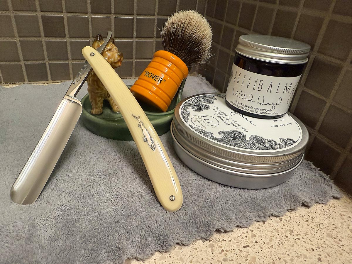 On a gray microfiber towel a straight razor with blade open about 45 degrees leans next to a bottle of aftershave and a jar of shave soap. A shave brush rests bristles-up in a green Wade ceramic pipe rest that features a brown Cairn Terrier.