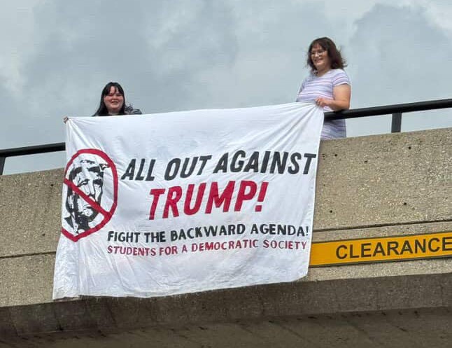 Two students stand on a bridge over a highway and drape a banner over the side of the bridge that says, 