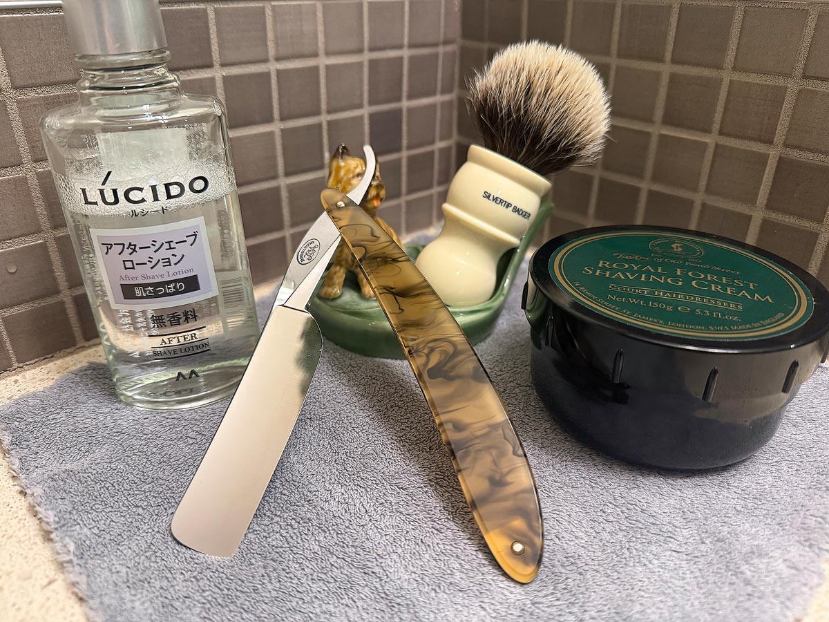 On a gray microfiber towel a straight razor with blade open about 45 degrees leans next to a bottle of aftershave and a jar of shave soap. A shave brush rests bristles-up in a green Wade ceramic pipe rest that features a brown Cairn Terrier.