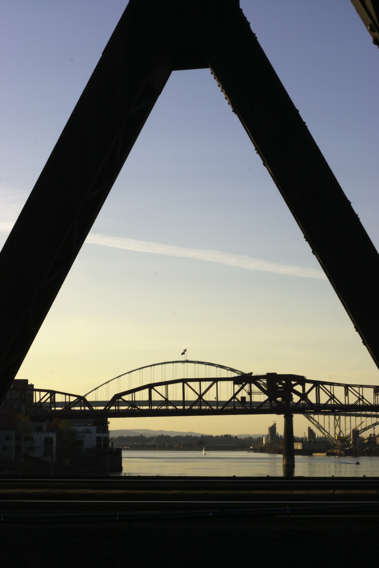 looking eastward down the willamette river at sunset, from between an angled support on the steel bridge. the broadway and fremont bridges sit above the river, sailboat enjoying the spring warmth.