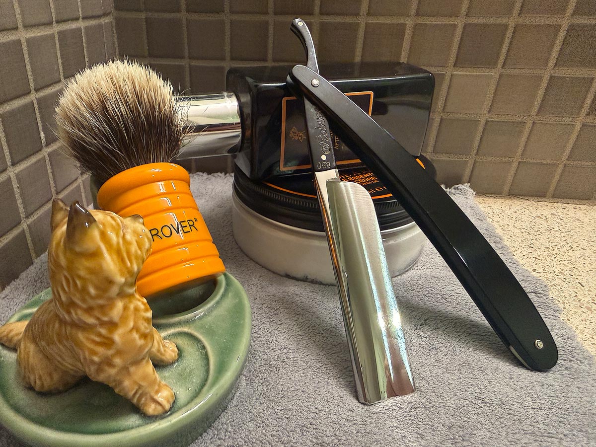 On a gray microfiber towel a straight razor with blade open about 45 degrees leans next to a bottle of aftershave and a jar of shave soap. A shave brush rests bristles-up in a green Wade ceramic pipe rest that features a brown Cairn Terrier.