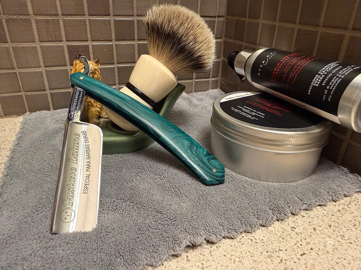 On a gray microfiber towel a straight razor with blade open about 45 degrees leans next to a bottle of aftershave and a jar of shave soap. A shave brush rests bristles-up in a green Wade ceramic pipe rest that features a brown Cairn Terrier.
