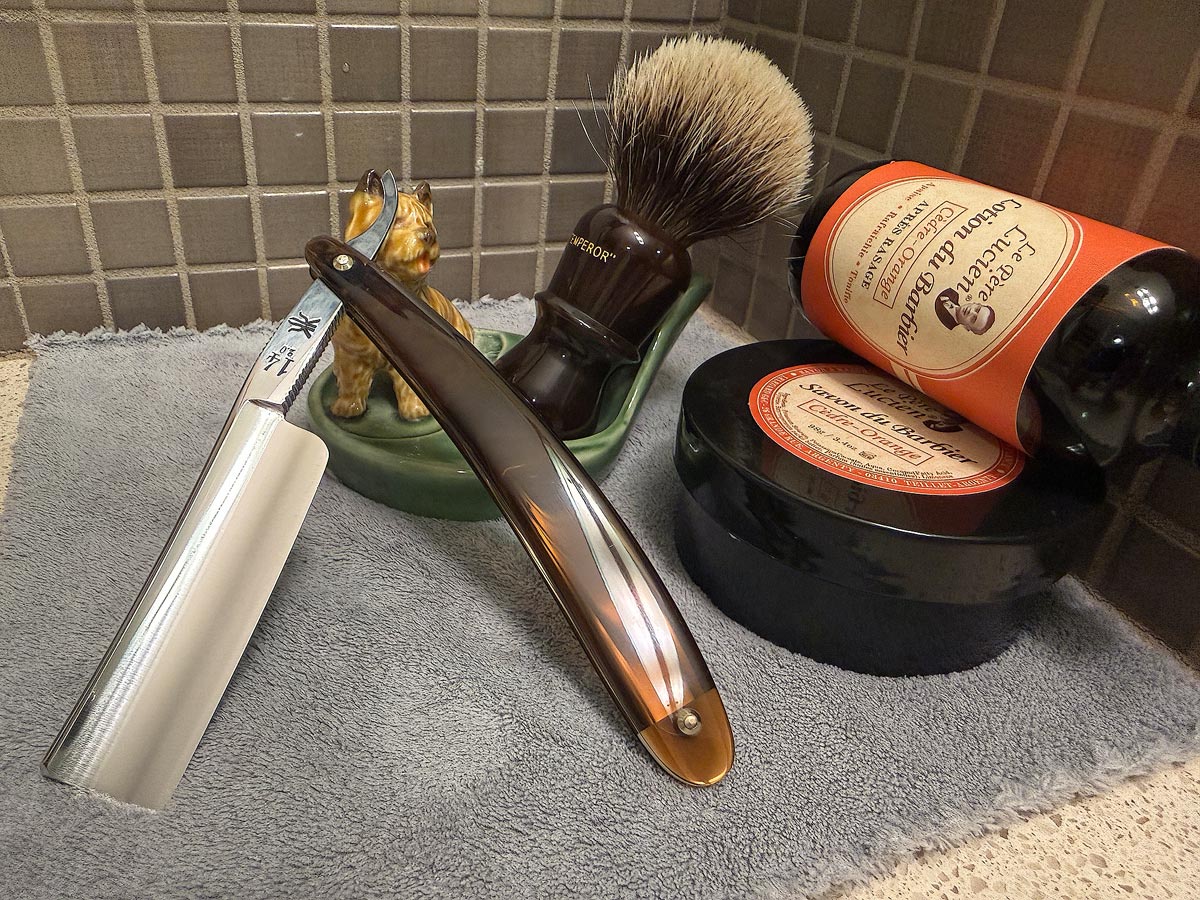 On a gray microfiber towel a straight razor with blade open about 45 degrees leans next to a bottle of aftershave and a jar of shave soap. A shave brush rests bristles-up in a green Wade ceramic pipe rest that features a brown Cairn Terrier.