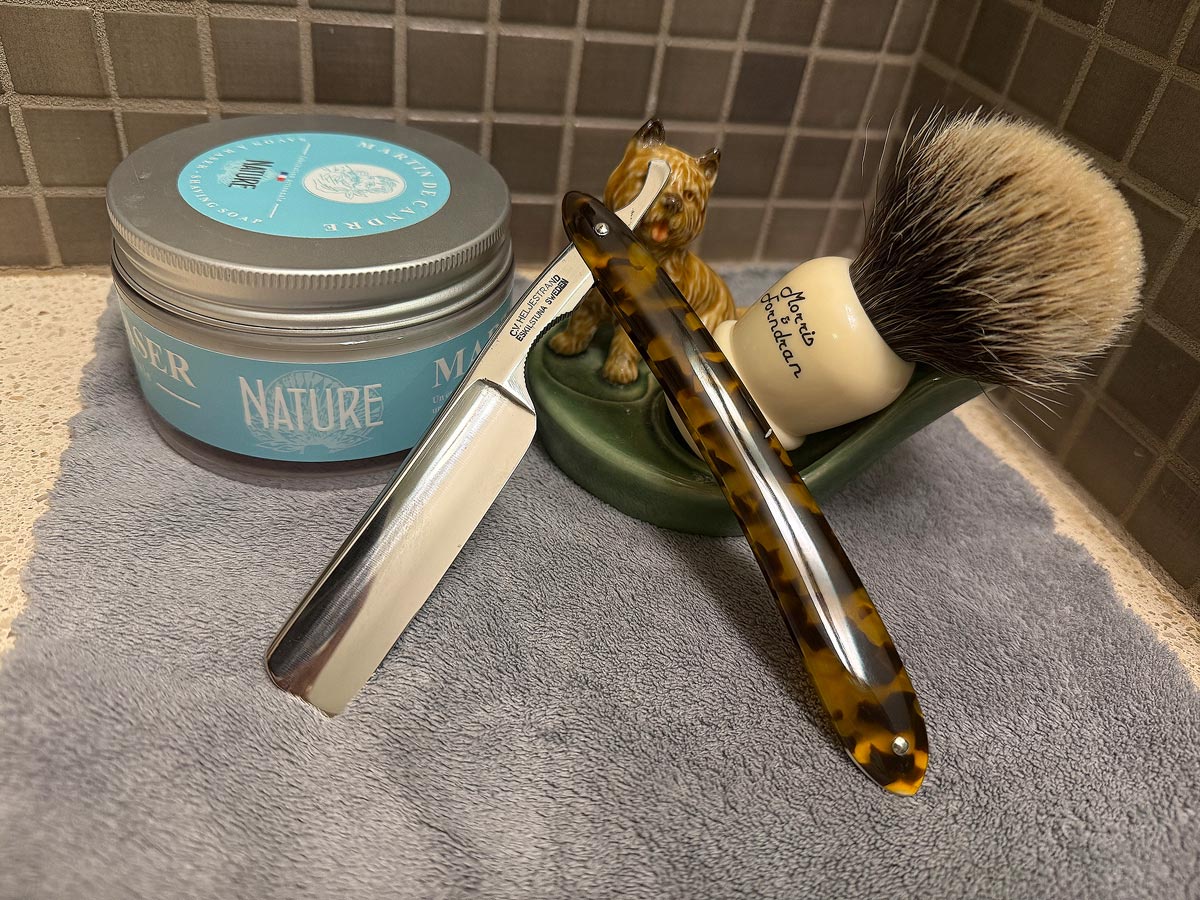 On a gray microfiber towel a straight razor with blade open about 45 degrees leans next to a bottle of aftershave and a jar of shave soap. A shave brush rests bristles-up in a green Wade ceramic pipe rest that features a brown Cairn Terrier.