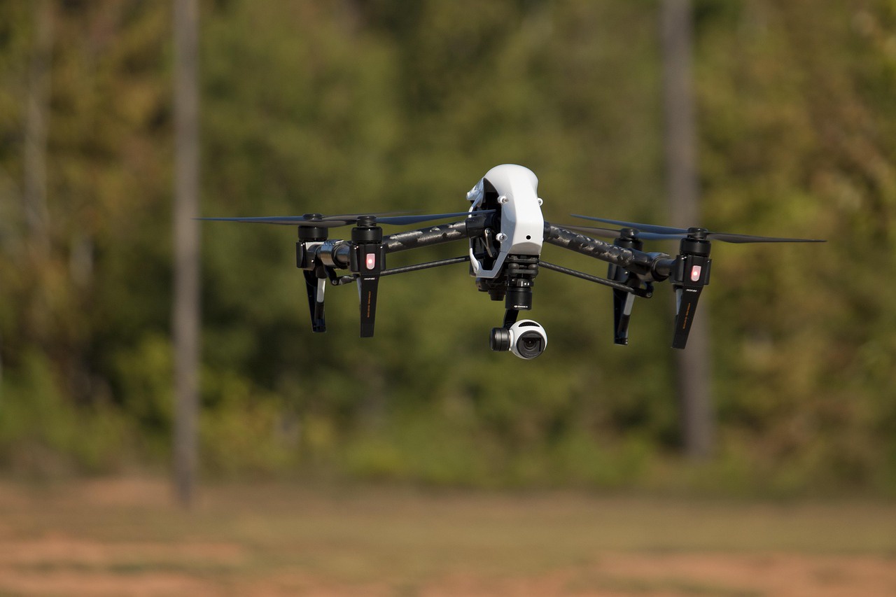 A white bodied quad copter floating in front of a line of trees. The copter's camera is pointed at the viewer.
