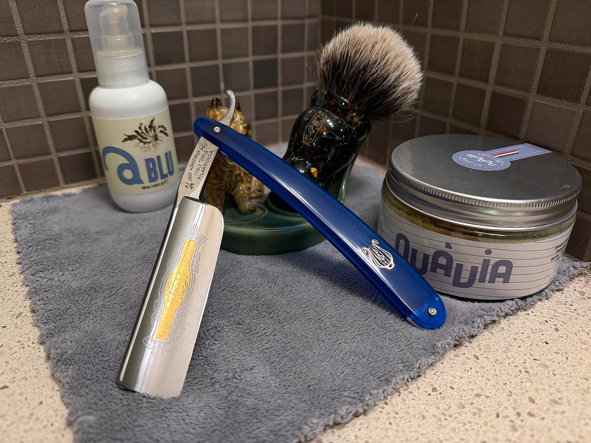 On a gray microfiber towel a straight razor with blade open about 45 degrees leans next to a bottle of aftershave and a jar of shave soap. A shave brush rests bristles-up in a green Wade ceramic pipe rest that features a brown Cairn Terrier.
