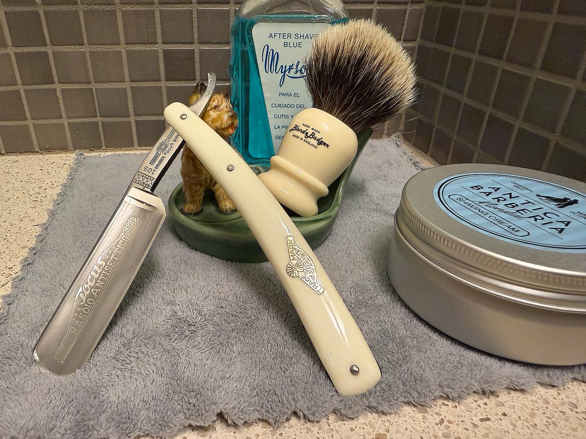 On a gray microfiber towel a straight razor with blade open about 45 degrees leans next to a bottle of aftershave and a jar of shave soap. A shave brush rests bristles-up in a green Wade ceramic pipe rest that features a brown Cairn Terrier.