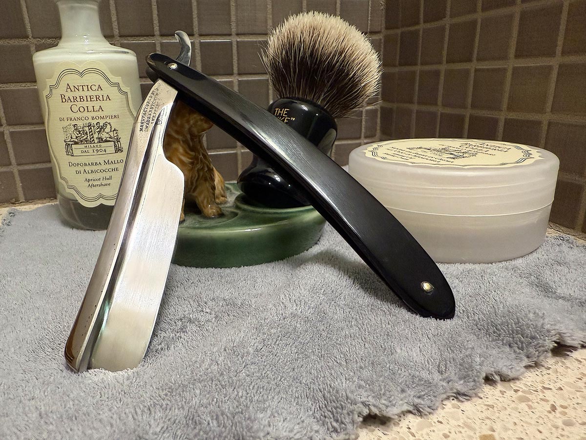 On a gray microfiber towel a straight razor with blade open about 45 degrees leans next to a bottle of aftershave and a jar of shave soap. A shave brush rests bristles-up in a green Wade ceramic pipe rest that features a brown Cairn Terrier.