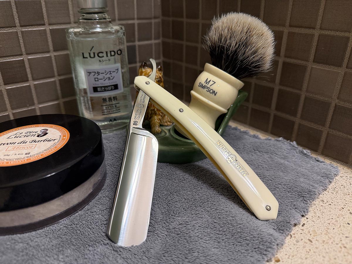 On a gray microfiber towel a straight razor with blade open about 45 degrees leans next to a bottle of aftershave and a jar of shave soap. A shave brush rests bristles-up in a green Wade ceramic pipe rest that features a brown Cairn Terrier.