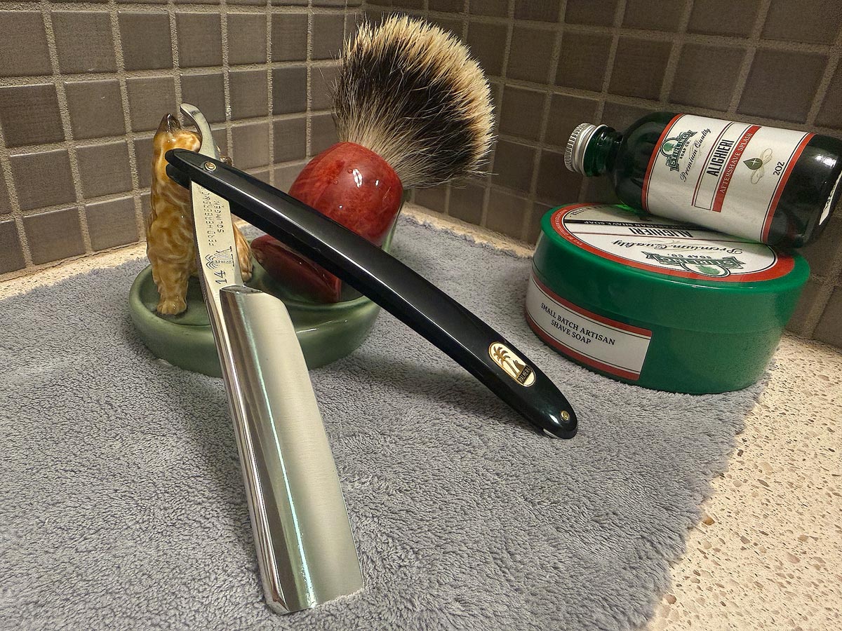 On a gray microfiber towel a straight razor with blade open about 45 degrees leans next to a bottle of aftershave and a jar of shave soap. A shave brush rests bristles-up in a green Wade ceramic pipe rest that features a brown Cairn Terrier.