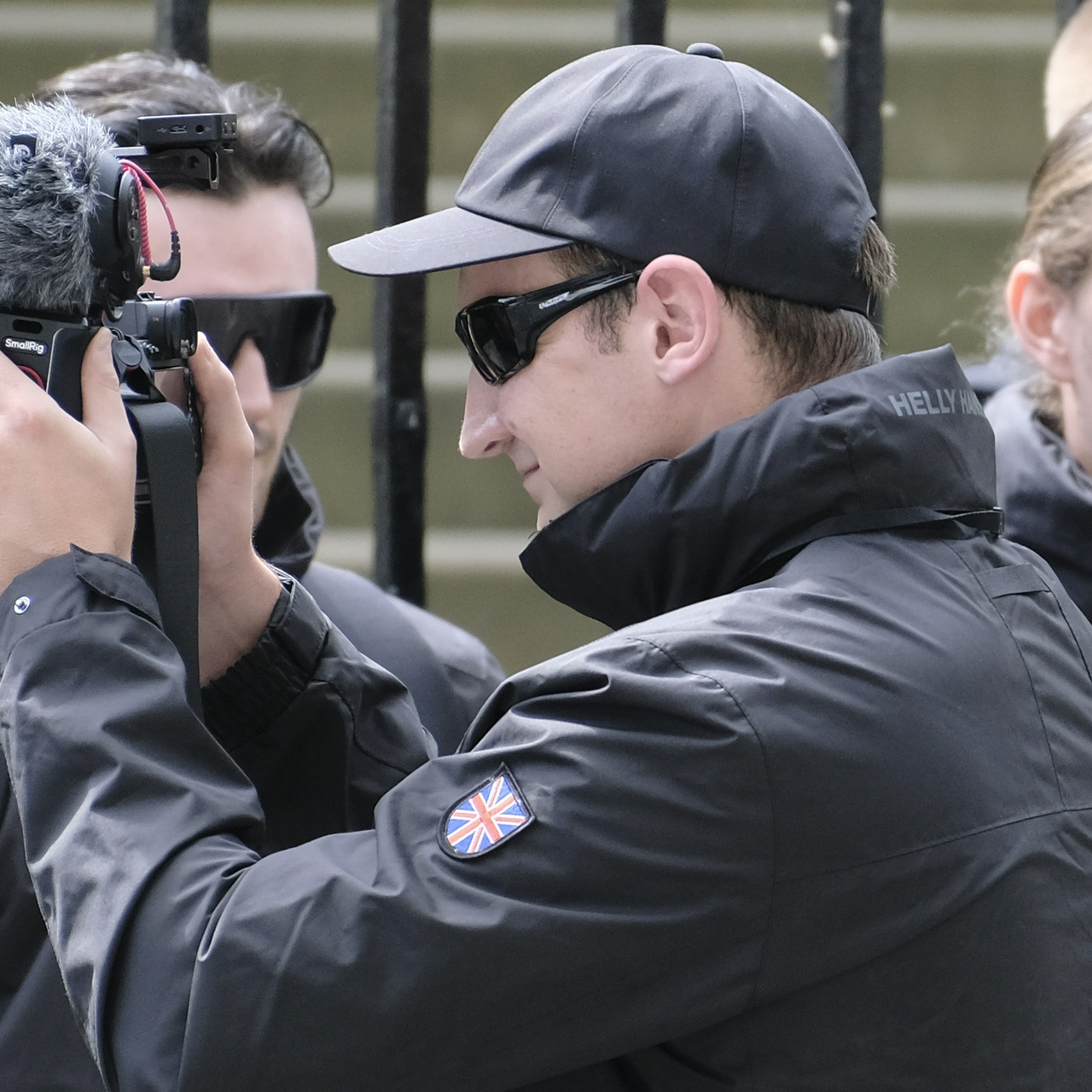 A tall young man with blonde hair sweats into his black cap as he grins at the video footage he's filming