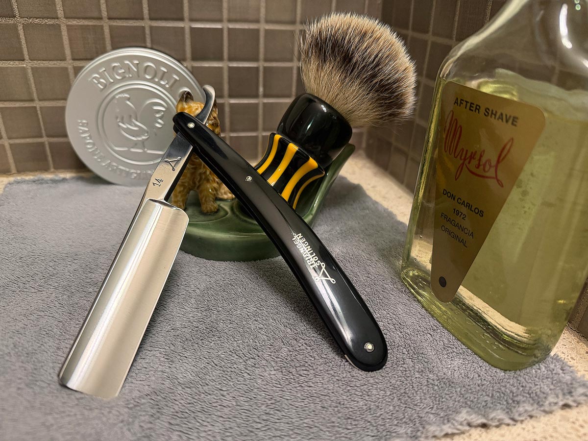 On a gray microfiber towel a straight razor with blade open about 45 degrees leans next to a bottle of aftershave and a jar of shave soap. A shave brush rests bristles-up in a green Wade ceramic pipe rest that features a brown Cairn Terrier.
