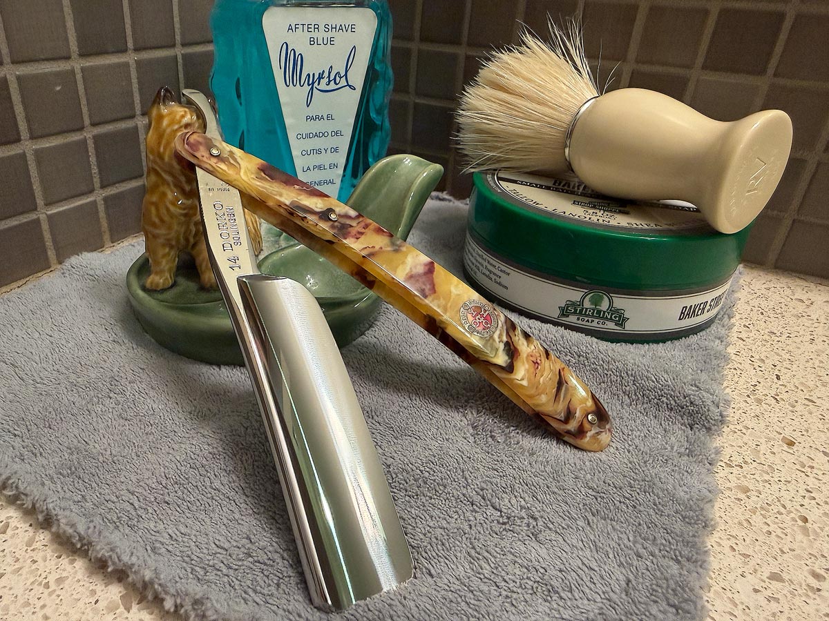 On a gray microfiber towel a straight razor with blade open about 45 degrees leans next to a bottle of aftershave and a jar of shave soap. A shave brush rests bristles-up in a green Wade ceramic pipe rest that features a brown Cairn Terrier.