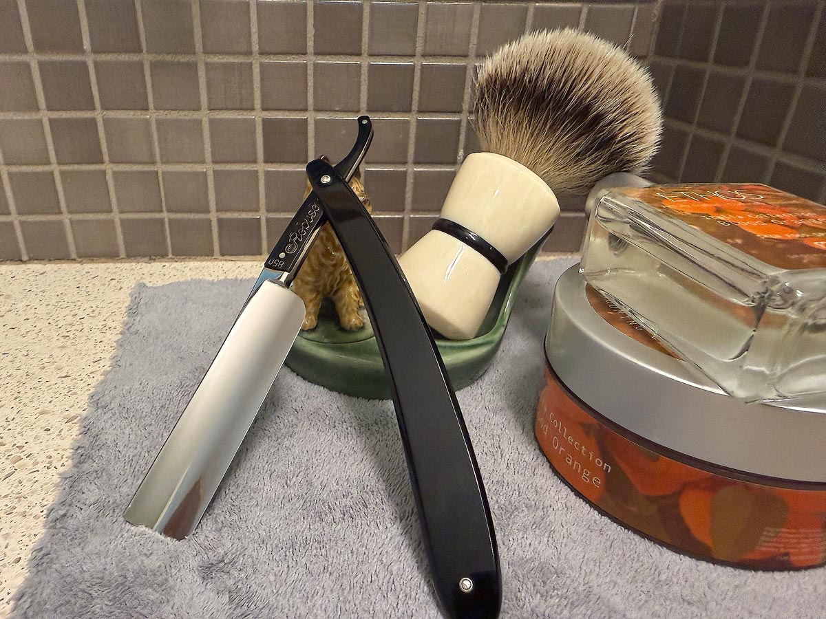 On a gray microfiber towel a straight razor with blade open about 45 degrees leans next to a bottle of aftershave and a jar of shave soap. A shave brush rests bristles-up in a green Wade ceramic pipe rest that features a brown Cairn Terrier.