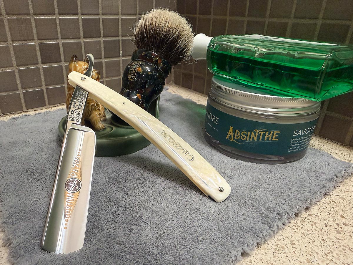 On a gray microfiber towel a straight razor with blade open about 45 degrees leans next to a bottle of aftershave and a jar of shave soap. A shave brush rests bristles-up in a green Wade ceramic pipe rest that features a brown Cairn Terrier.