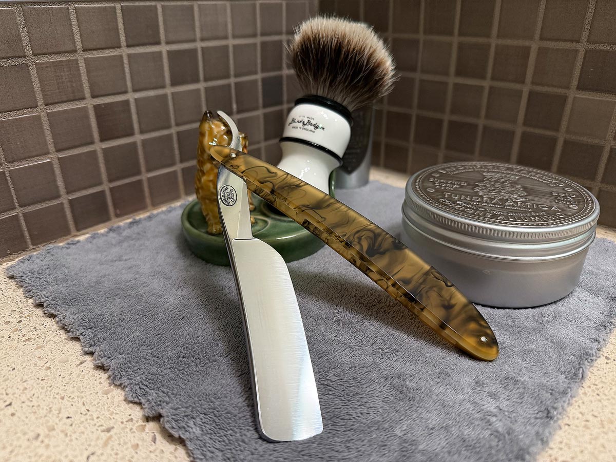 On a gray microfiber towel a straight razor with blade open about 45 degrees leans next to a bottle of aftershave and a jar of shave soap. A shave brush rests bristles-up in a green Wade ceramic pipe rest that features a brown Cairn Terrier.
