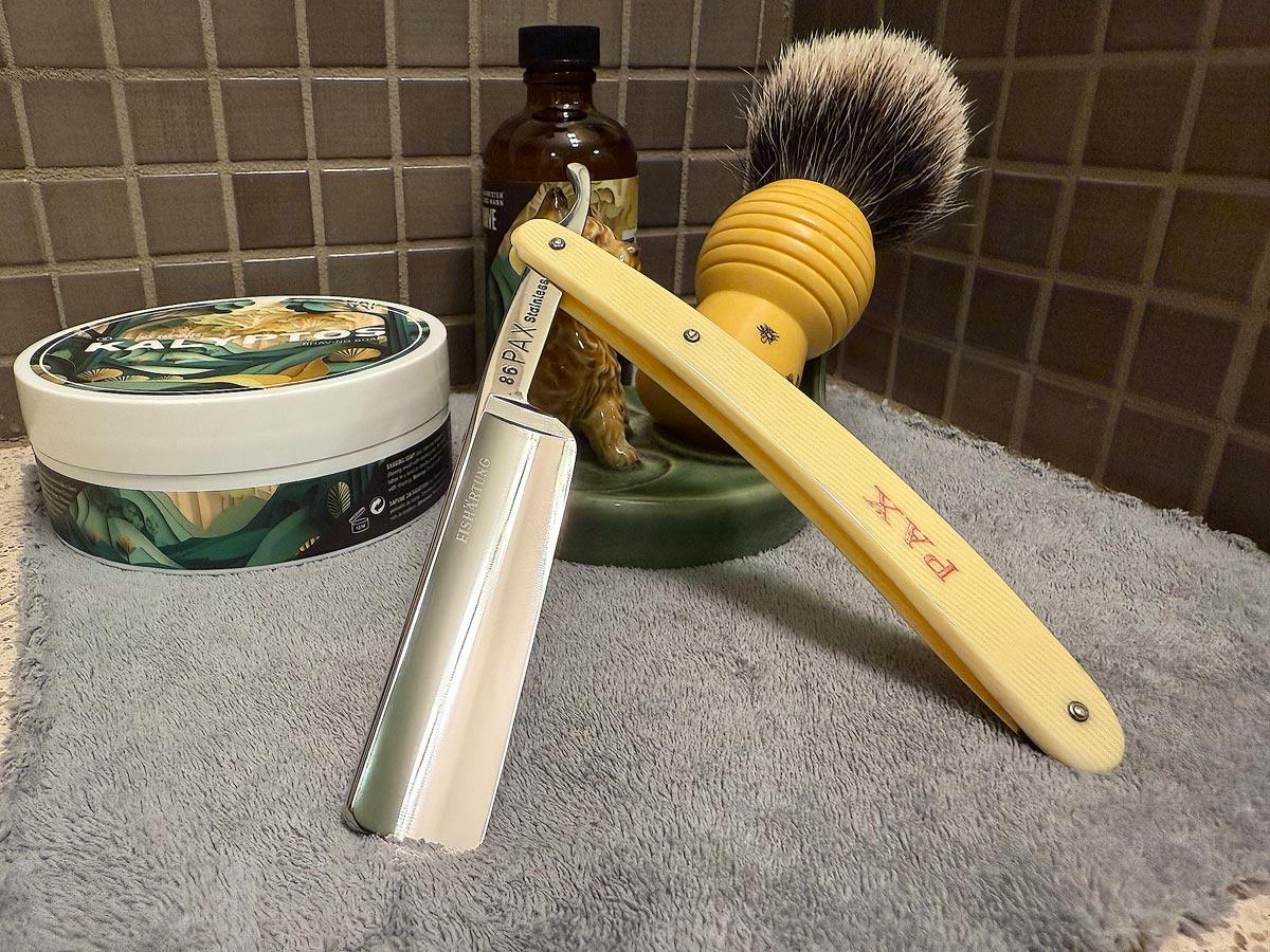 On a gray microfiber towel a straight razor with blade open about 45 degrees leans next to a bottle of aftershave and a jar of shave soap. A shave brush rests bristles-up in a green Wade ceramic pipe rest that features a brown Cairn Terrier.