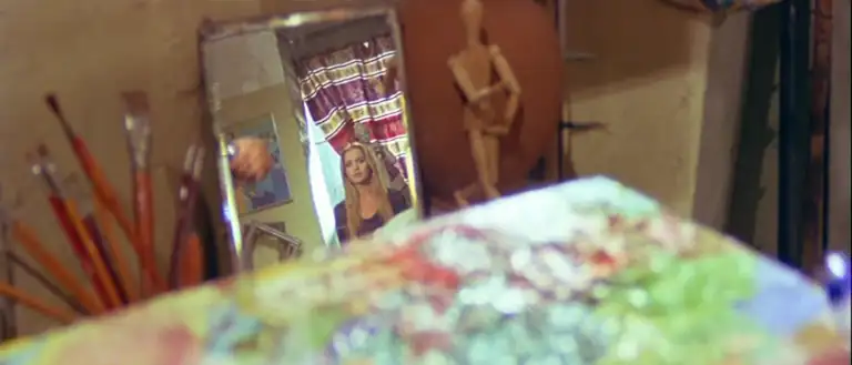 Loredana shown in a mirror in a makeshift painter's studio. She has long, blonde hair and wears a long sleeved purple top. There is a small, wood mannikin posed beside the mirror. The other side of the mirror shows pain brushes in a container. A square of paint is in the foreground of the image, operating as a paint palette.