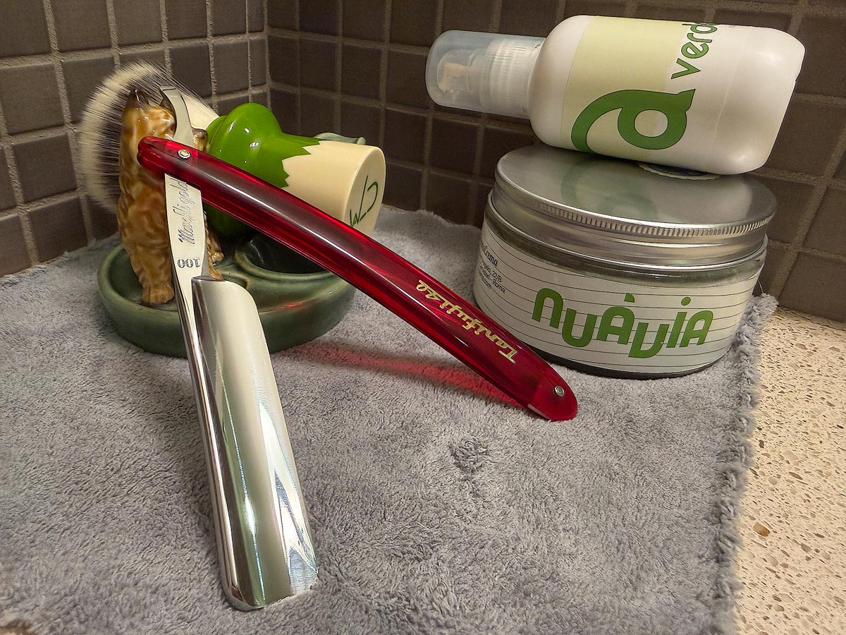 On a gray microfiber towel a straight razor with blade open about 45 degrees leans next to a bottle of aftershave and a jar of shave soap. A shave brush rests bristles-up in a green Wade ceramic pipe rest that features a brown Cairn Terrier.