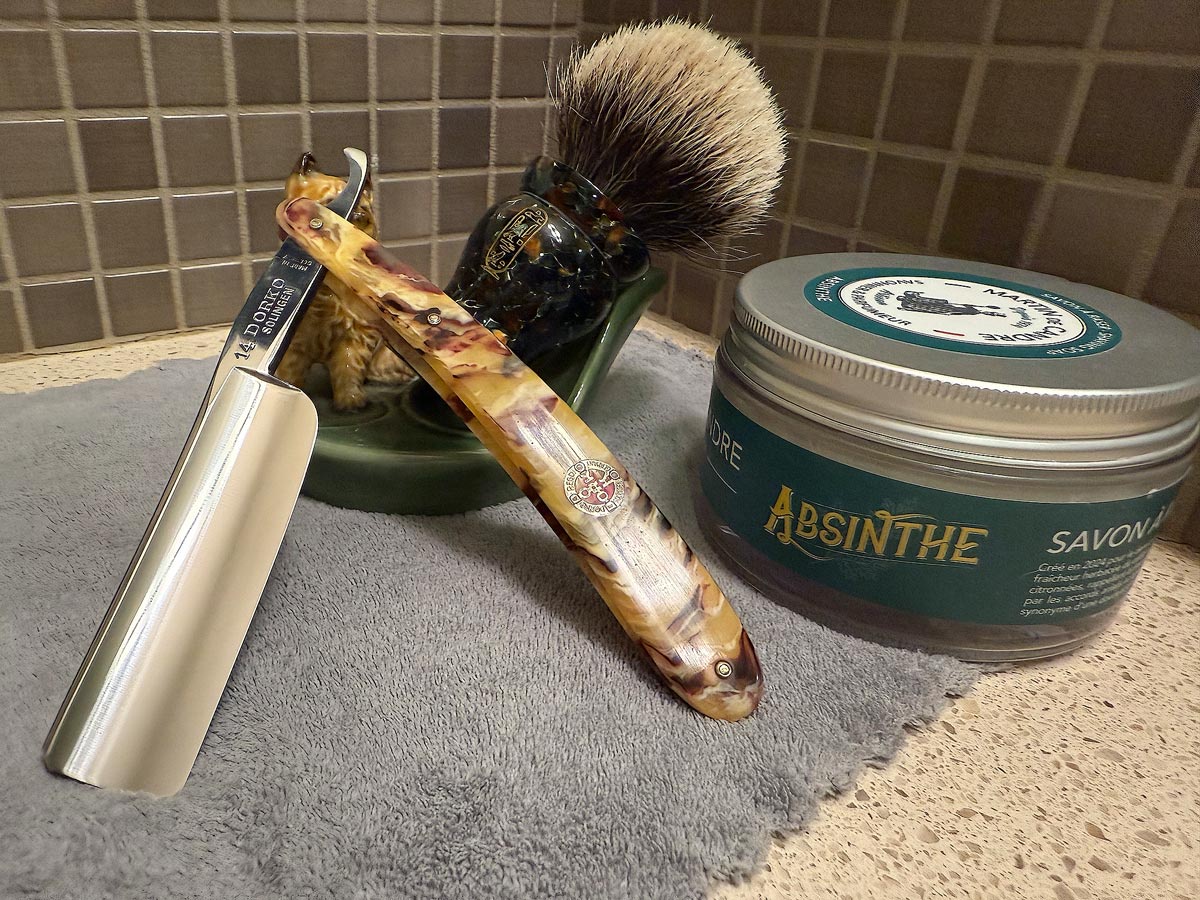 On a gray microfiber towel a straight razor with blade open about 45 degrees leans next to a bottle of aftershave and a jar of shave soap. A shave brush rests bristles-up in a green Wade ceramic pipe rest that features a brown Cairn Terrier.