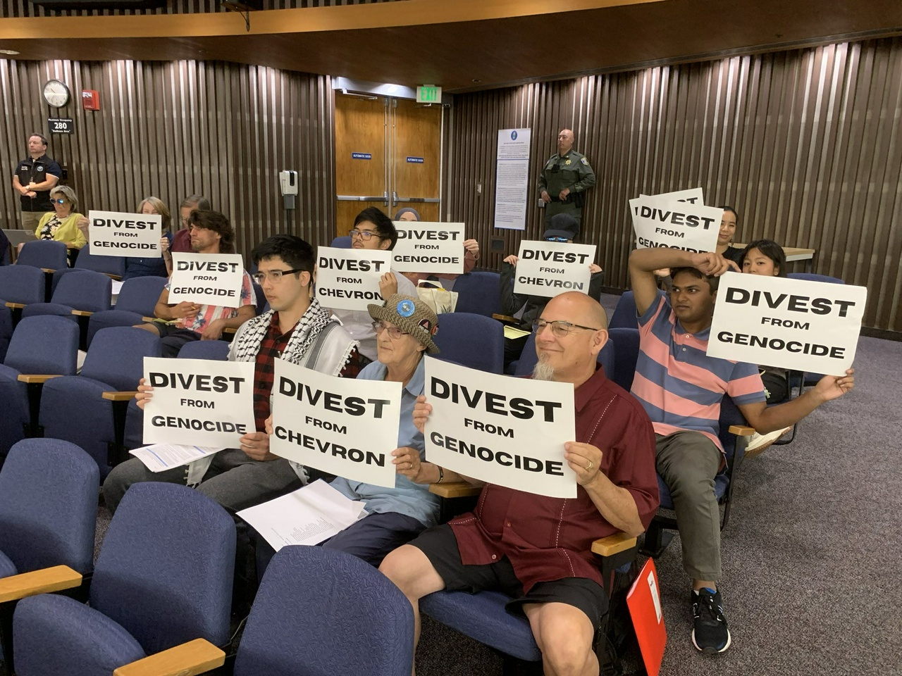 A group of protesters sit in a city subcommittee meeting and hold signs that say, 