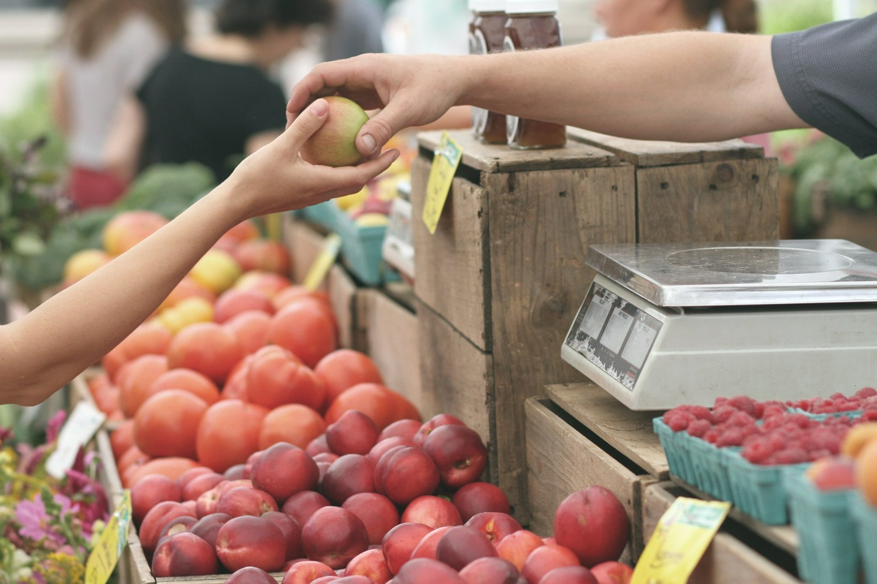 One person hands another an apple at a fruit stand. A scale is visible in the foreground, suggesting an exchange of money.