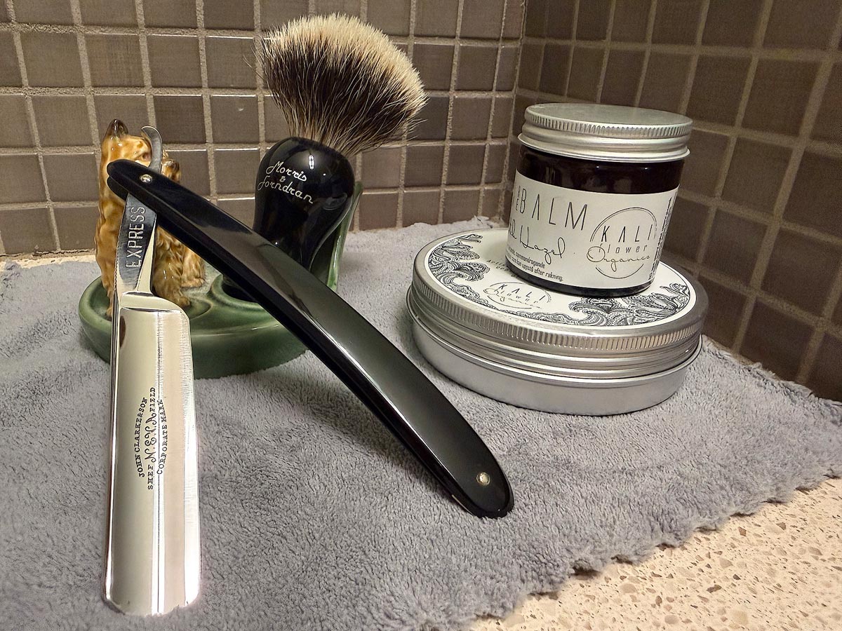 On a gray microfiber towel a straight razor with blade open about 45 degrees leans next to a bottle of aftershave and a jar of shave soap. A shave brush rests bristles-up in a green Wade ceramic pipe rest that features a brown Cairn Terrier.