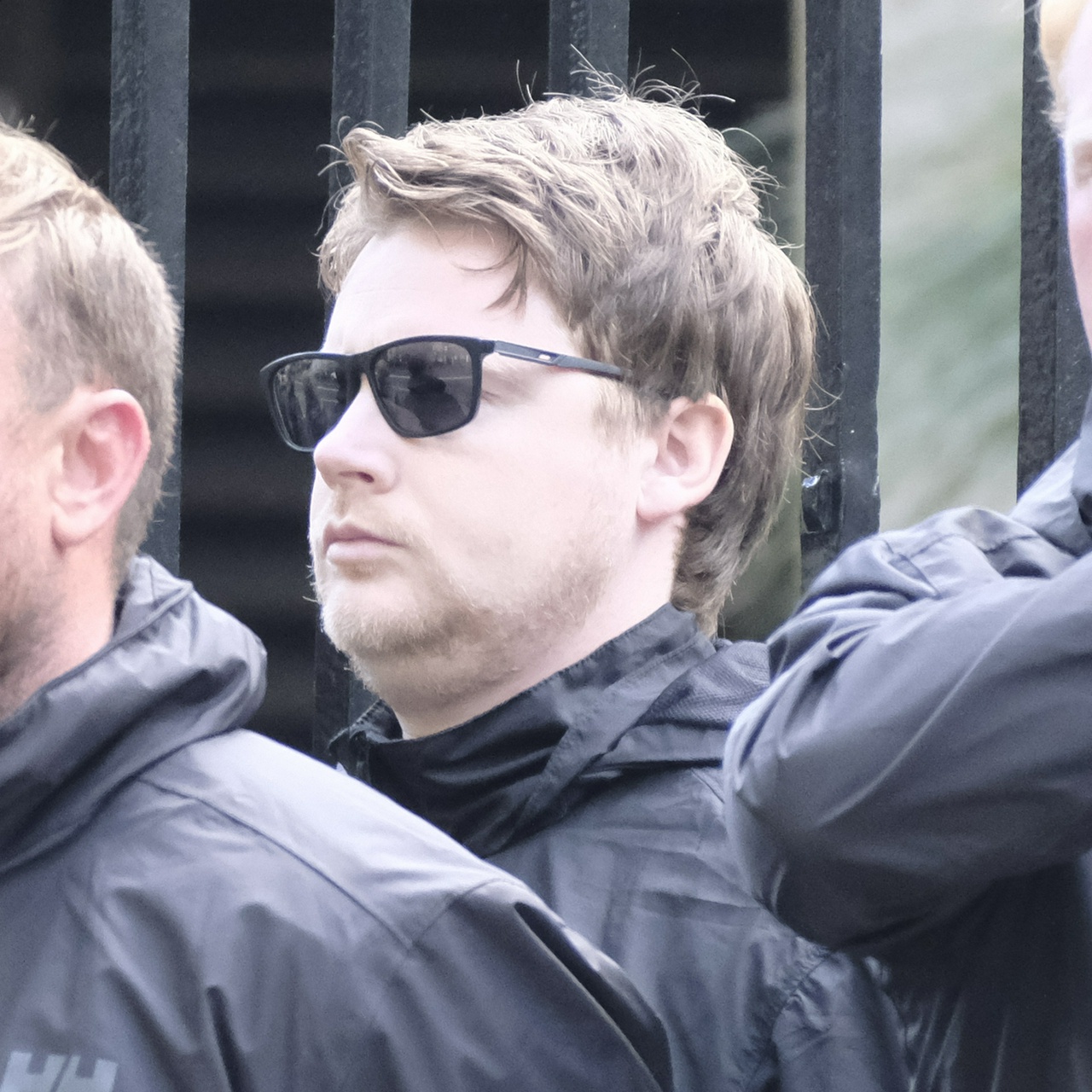 A double chinned white man with tousled, mousy brown hair and black sunglasses
