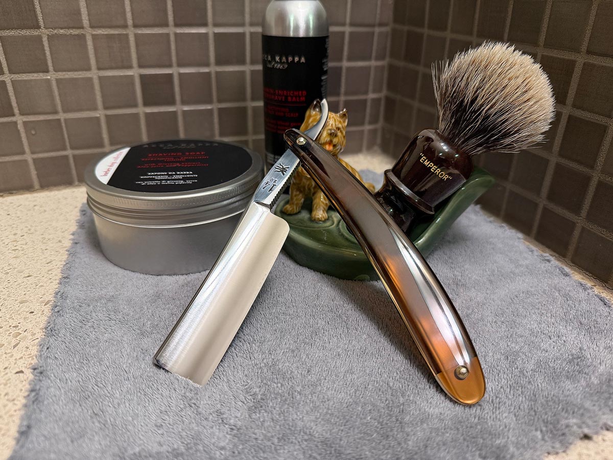 On a gray microfiber towel a straight razor with blade open about 45 degrees leans next to a bottle of aftershave and a jar of shave soap. A shave brush rests bristles-up in a green Wade ceramic pipe rest that features a brown Cairn Terrier.