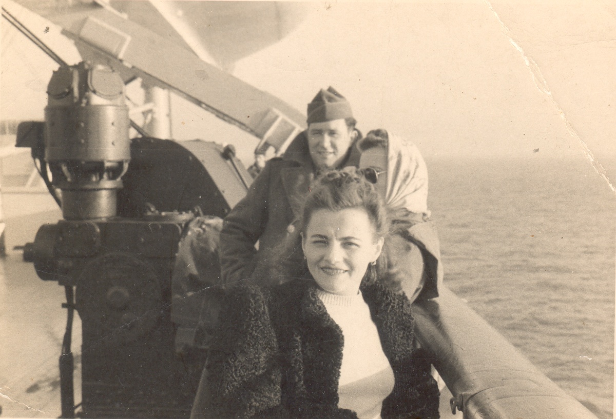 A vintage black and white photo of a smiling woman in a fur coat and a man in military uniform, both on the deck of a ship. The man is partially obscured by the woman who is seated in the foreground. Behind them, part of a naval anti-aircraft gun is visible, suggesting a wartime setting. The ocean and a hazy sky form the backdrop, enhancing the historical feel of the image.