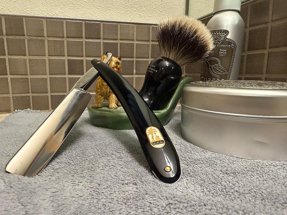 On a gray microfiber towel a straight razor with blade open about 45 degrees leans next to a bottle of aftershave and a jar of shave soap. A shave brush rests bristles-up in a green Wade ceramic pipe rest that features a brown Cairn Terrier.