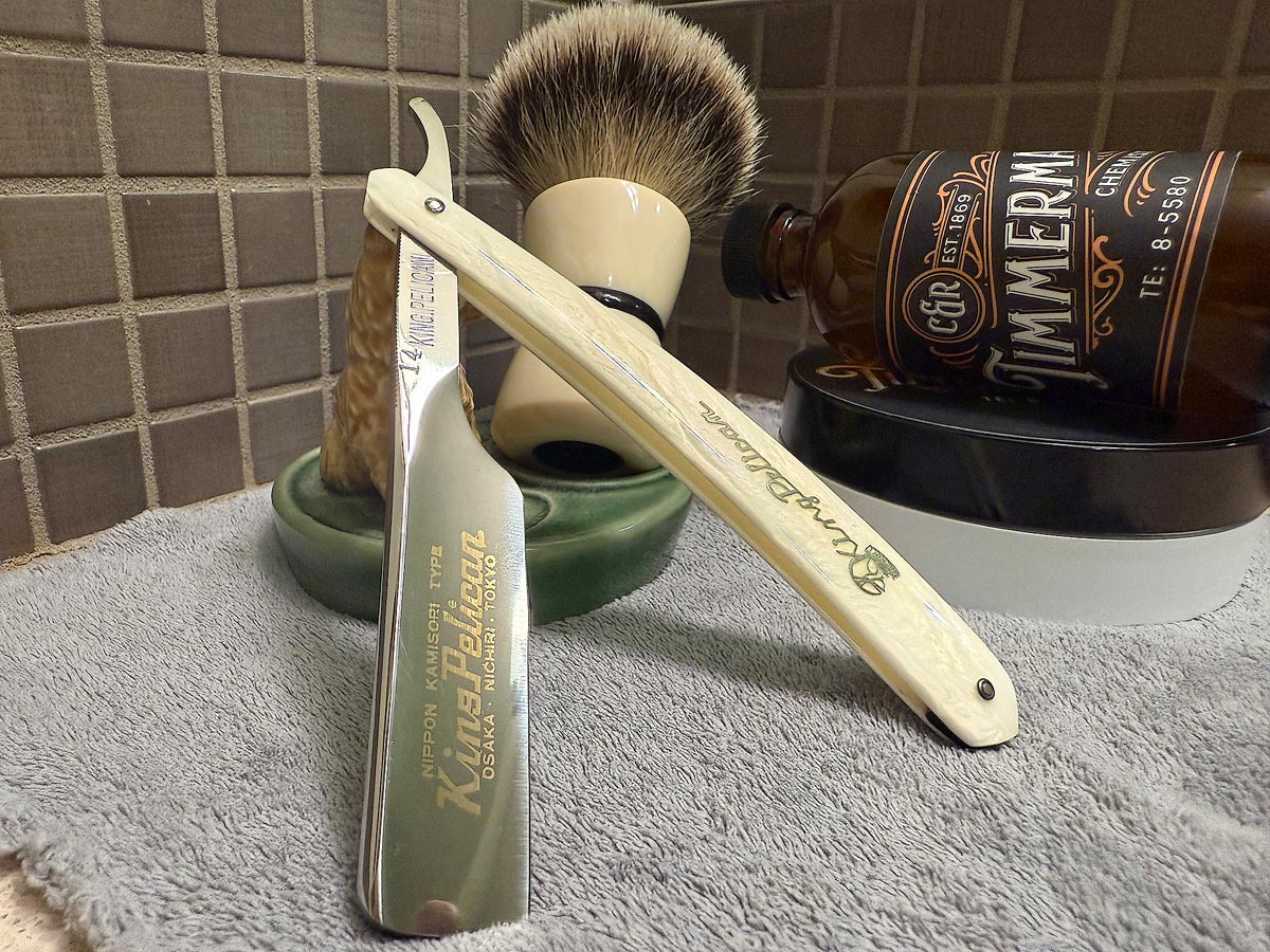 On a gray microfiber towel a straight razor with blade open about 45 degrees leans next to a bottle of aftershave and a jar of shave soap. A shave brush rests bristles-up in a green Wade ceramic pipe rest that features a brown Cairn Terrier.