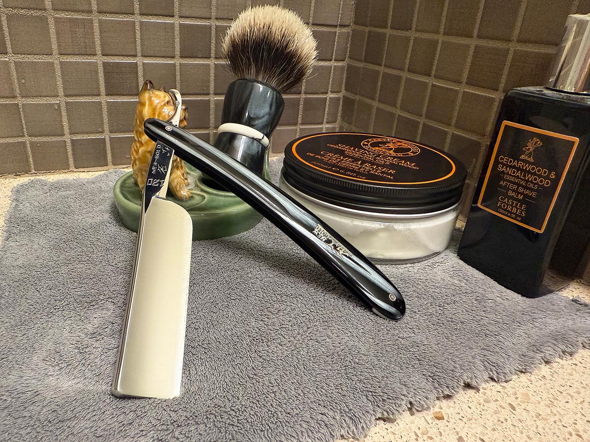 On a gray microfiber towel a straight razor with blade open about 45 degrees leans next to a bottle of aftershave and a jar of shave soap. A shave brush rests bristles-up in a green Wade ceramic pipe rest that features a brown Cairn Terrier.