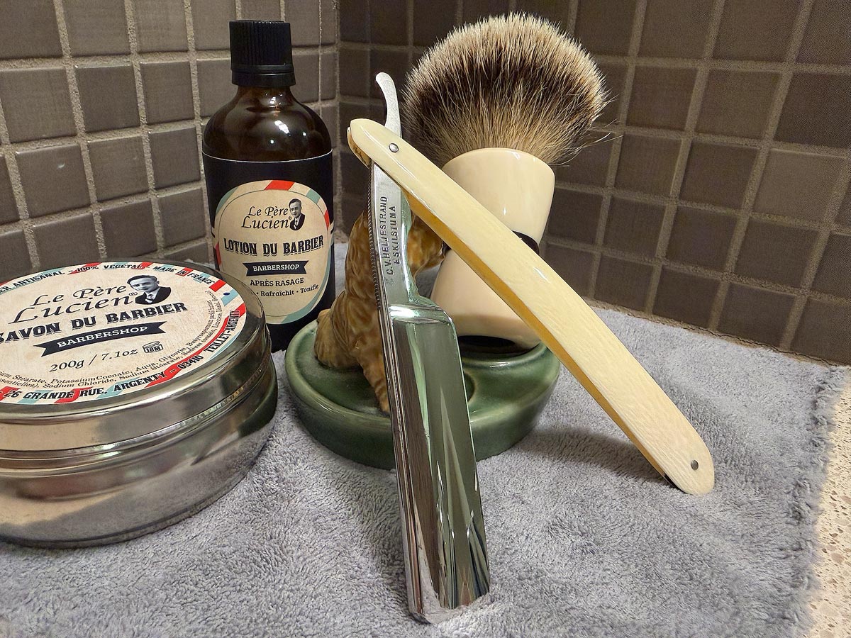 On a gray microfiber towel a straight razor with blade open about 45 degrees leans next to a bottle of aftershave and a jar of shave soap. A shave brush rests bristles-up in a green Wade ceramic pipe rest that features a brown Cairn Terrier.