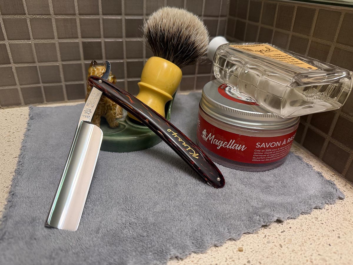 On a gray microfiber towel a straight razor with blade open about 45 degrees leans next to a bottle of aftershave and a jar of shave soap. A shave brush rests bristles-up in a green Wade ceramic pipe rest that features a brown Cairn Terrier.