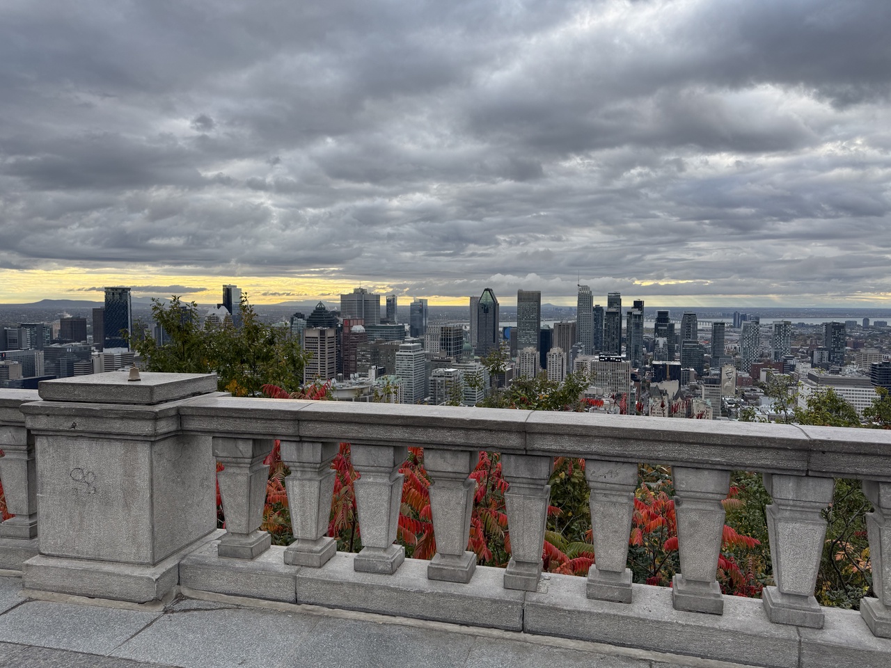 A view over downtown Montréal from a stone balustrade at the lookout, under heavy grey clouds with a thin band of yellow light along the horizon. Red sumac leaves show through the balusters in the foreground.