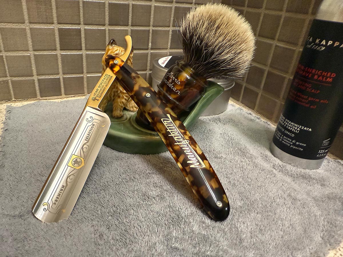 On a gray microfiber towel a straight razor with blade open about 45 degrees leans next to a bottle of aftershave and a jar of shave soap. A shave brush rests bristles-up in a green Wade ceramic pipe rest that features a brown Cairn Terrier.
