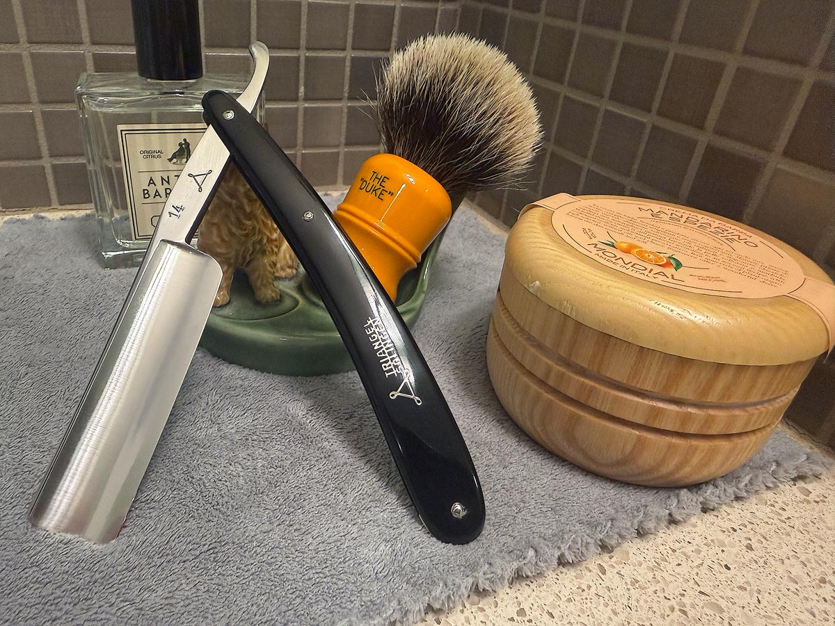 On a gray microfiber towel a straight razor with blade open about 45 degrees leans next to a bottle of aftershave and a jar of shave soap. A shave brush rests bristles-up in a green Wade ceramic pipe rest that features a brown Cairn Terrier.