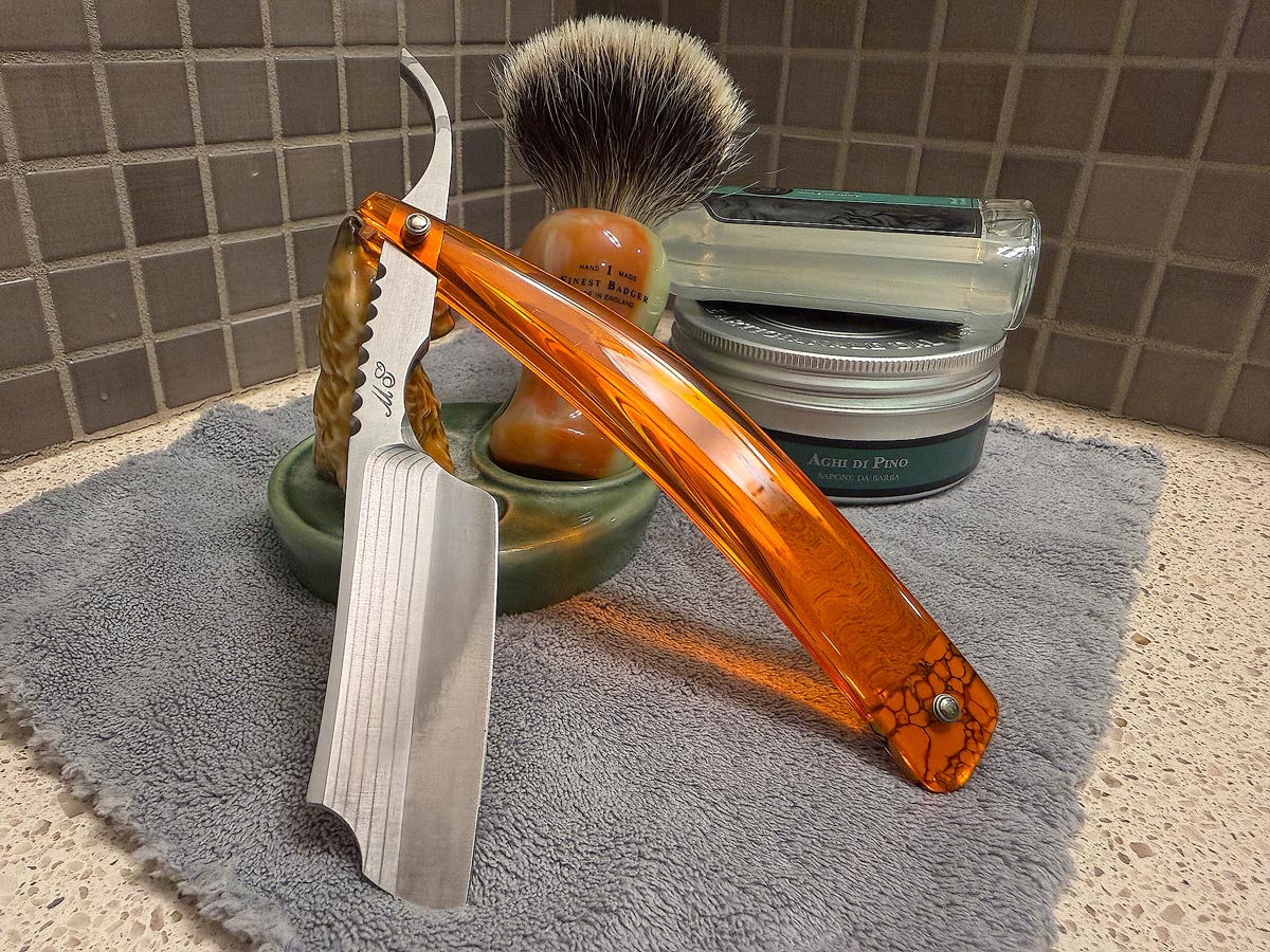 On a gray microfiber towel a straight razor with blade open about 45 degrees leans next to a bottle of aftershave and a jar of shave soap. A shave brush rests bristles-up in a green Wade ceramic pipe rest that features a brown Cairn Terrier.