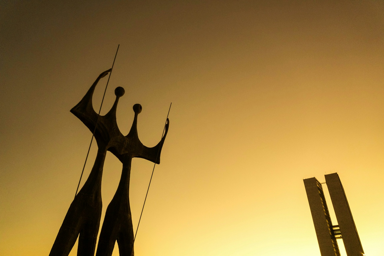 Two human-like statues in front of an iconic building, with the sky lit up all yellow-orange