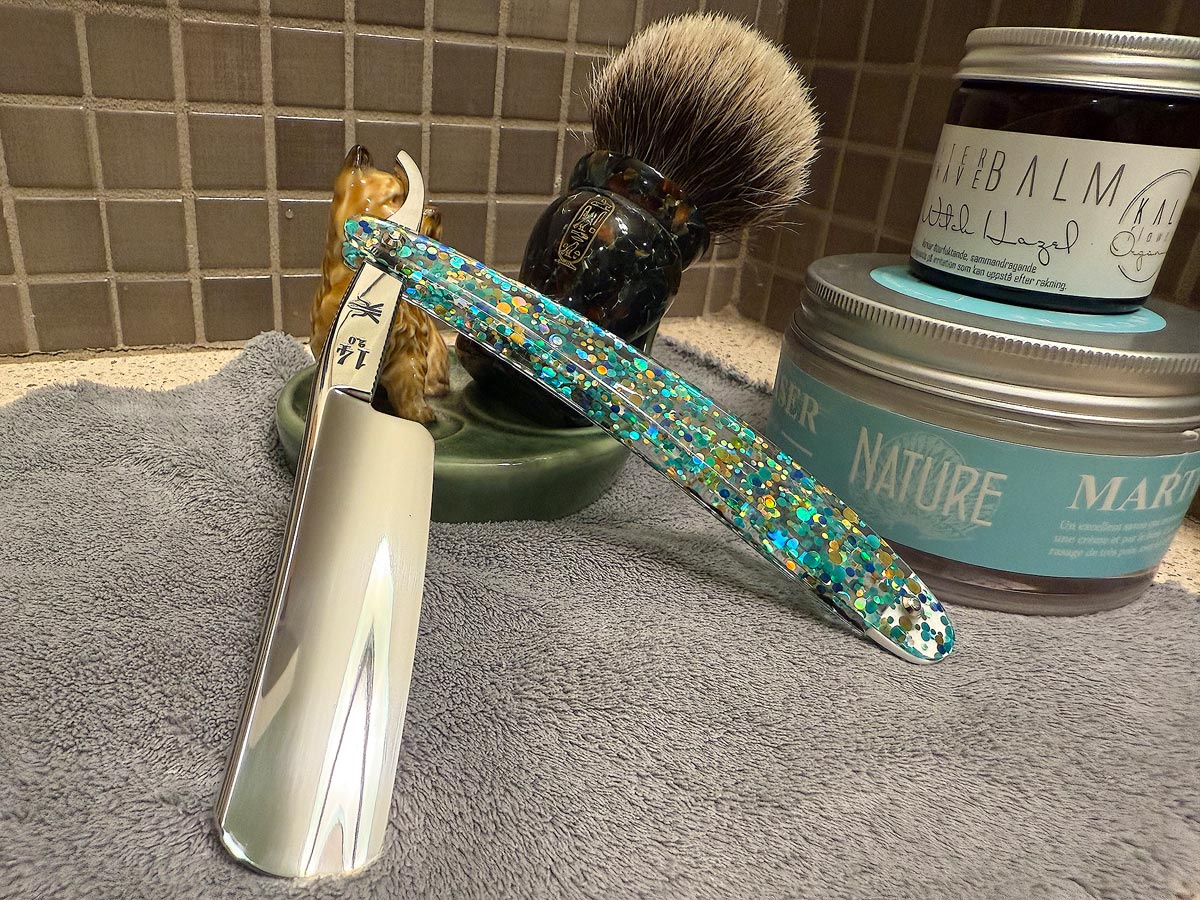 On a gray microfiber towel a straight razor with blade open about 45 degrees leans next to a bottle of aftershave and a jar of shave soap. A shave brush rests bristles-up in a green Wade ceramic pipe rest that features a brown Cairn Terrier.