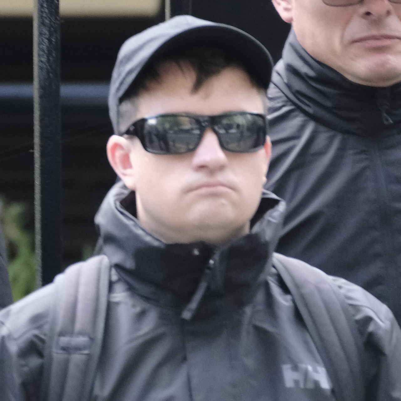 A short broad man with a wide nose and a perma-frown in a black cap and sunnies