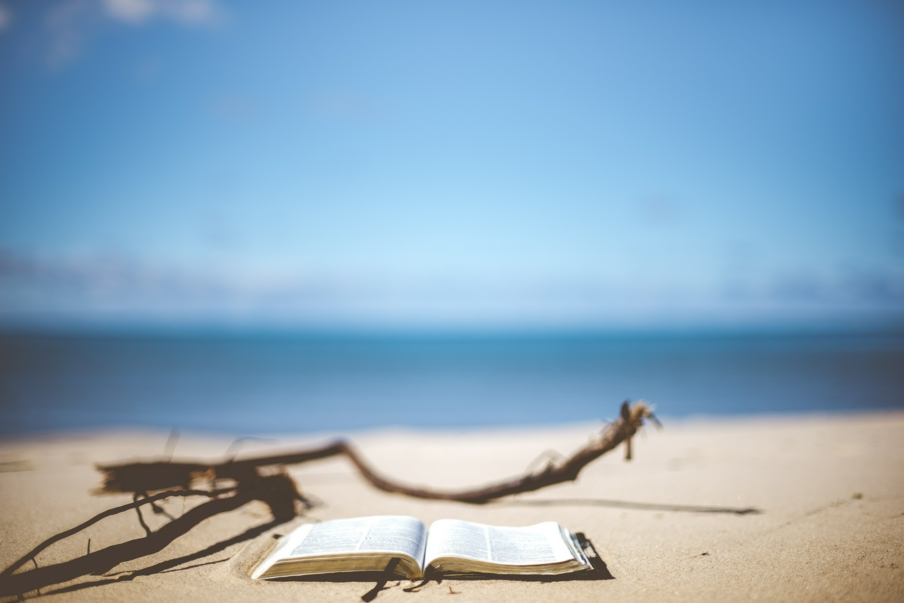 An open book lying on a sandy beach, with the water and sky visible in the background. No people are pictured nearby.