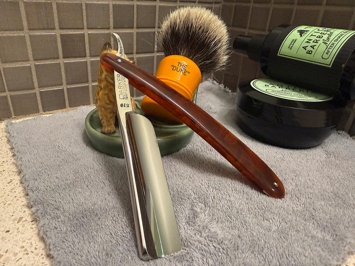 On a gray microfiber towel a straight razor with blade open about 45 degrees leans next to a bottle of aftershave and a jar of shave soap. A shave brush rests bristles-up in a green Wade ceramic pipe rest that features a brown Cairn Terrier.