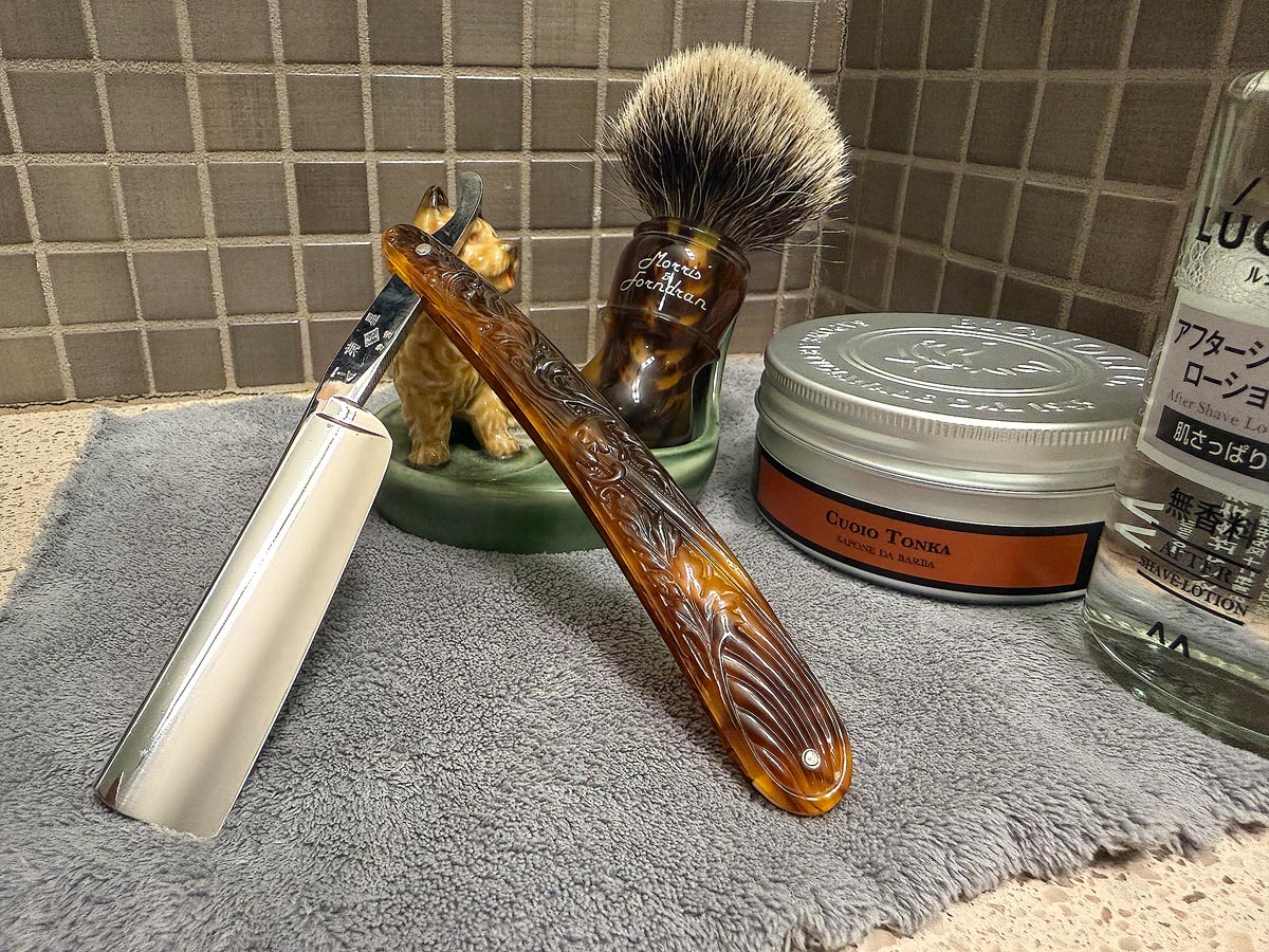 On a gray microfiber towel a straight razor with blade open about 45 degrees leans next to a bottle of aftershave and a jar of shave soap. A shave brush rests bristles-up in a green Wade ceramic pipe rest that features a brown Cairn Terrier.