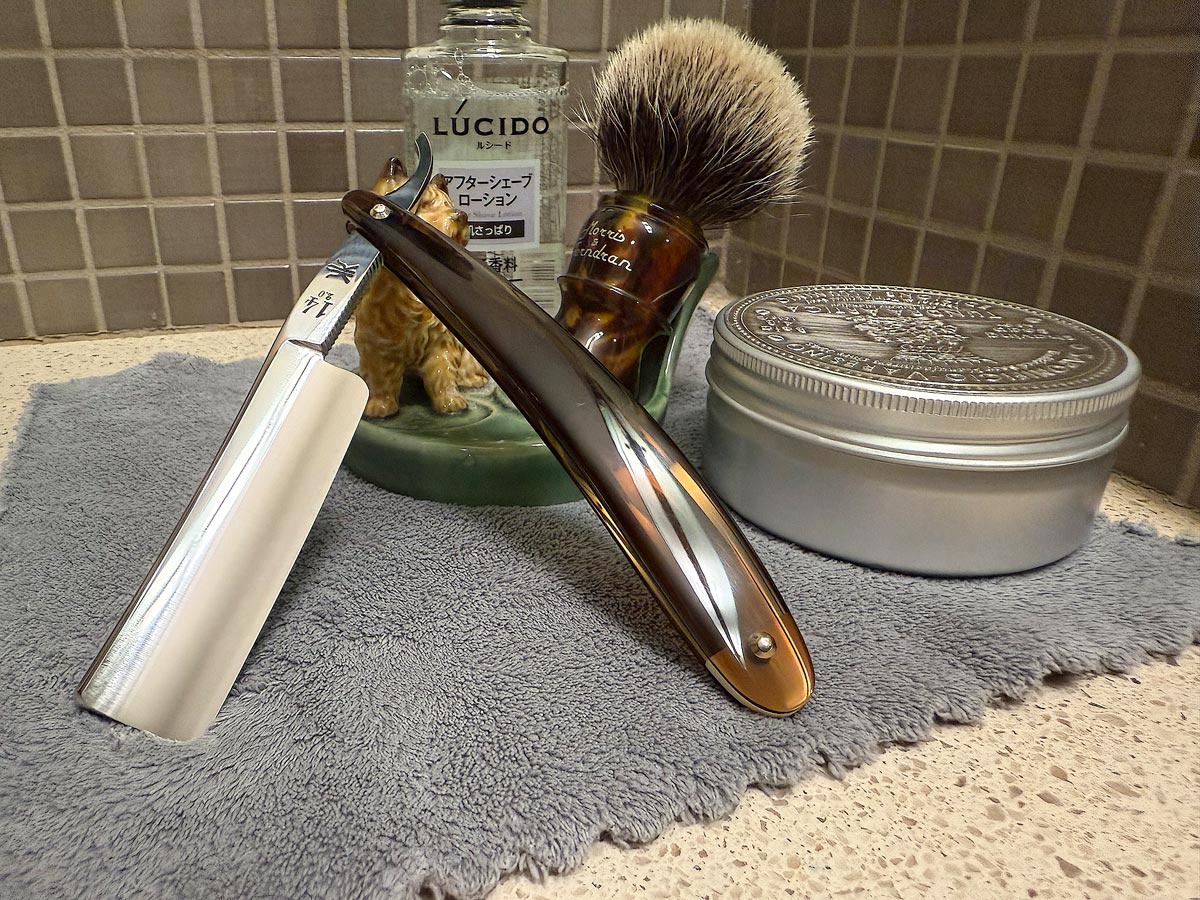 On a gray microfiber towel a straight razor with blade open about 45 degrees leans next to a bottle of aftershave and a jar of shave soap. A shave brush rests bristles-up in a green Wade ceramic pipe rest that features a brown Cairn Terrier.