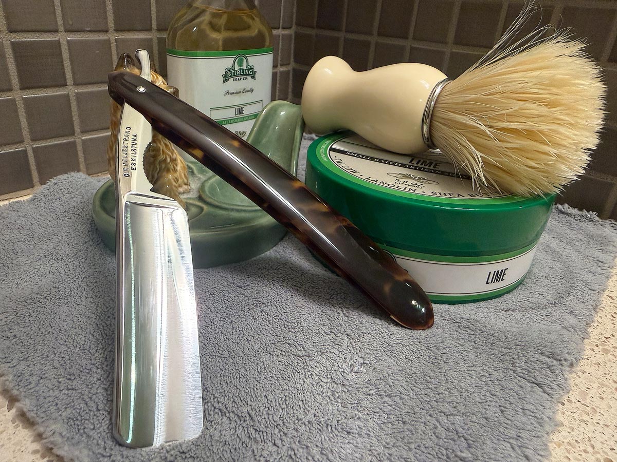 On a gray microfiber towel a straight razor with blade open about 45 degrees leans next to a bottle of aftershave and a jar of shave soap. A shave brush rests bristles-up in a green Wade ceramic pipe rest that features a brown Cairn Terrier.