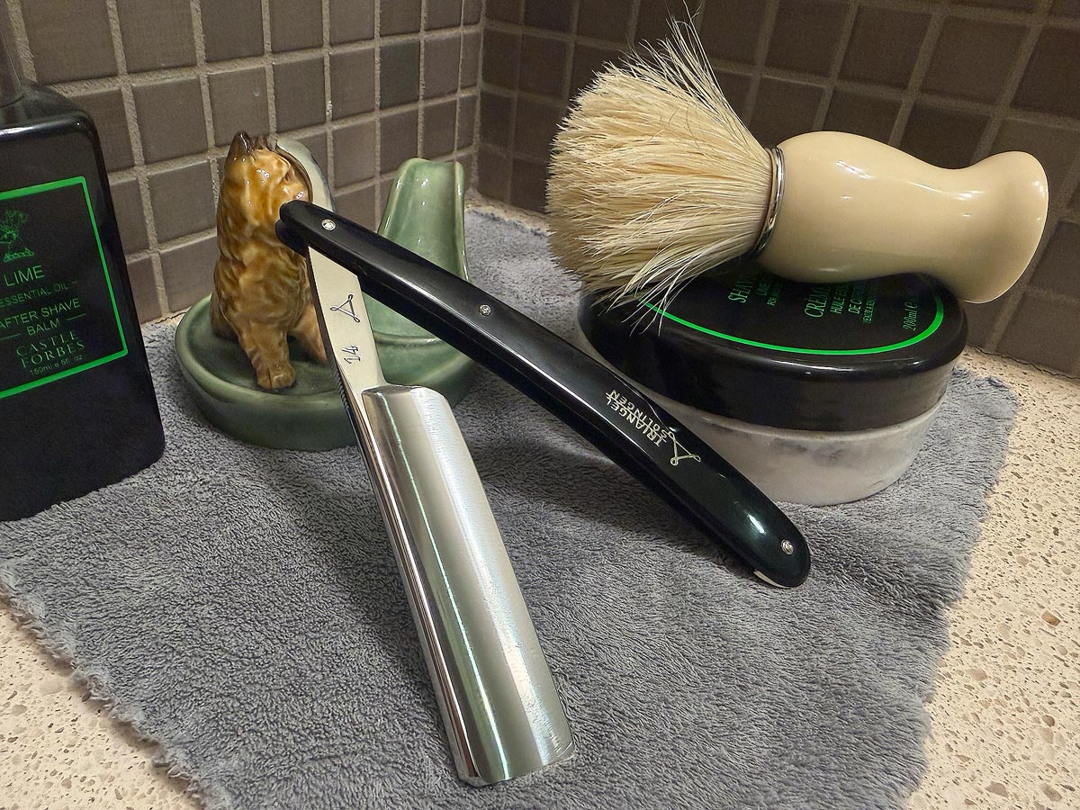On a gray microfiber towel a straight razor with blade open about 45 degrees leans next to a bottle of aftershave and a jar of shave soap. A shave brush rests bristles-up in a green Wade ceramic pipe rest that features a brown Cairn Terrier.