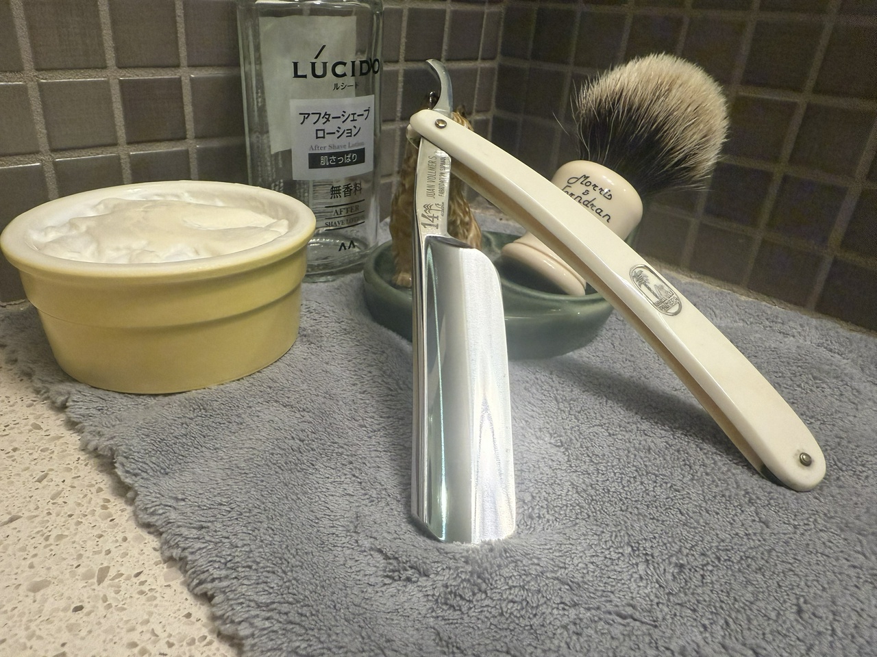 On a gray microfiber towel a straight razor with blade open about 45 degrees leans next to a bottle of aftershave and a jar of shave soap. A shave brush rests bristles-up in a green Wade ceramic pipe rest that features a brown Cairn Terrier.
