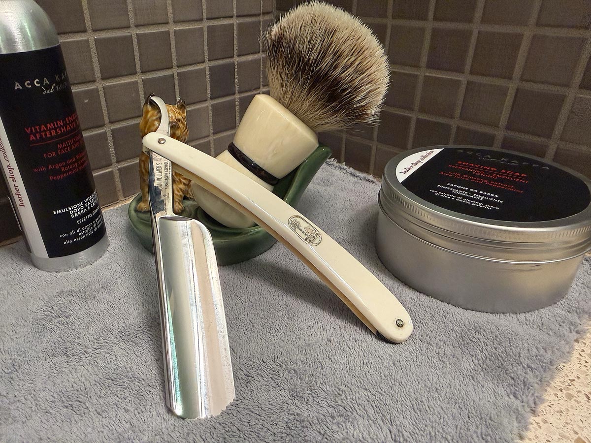 On a gray microfiber towel a straight razor with blade open about 45 degrees leans next to a bottle of aftershave and a jar of shave soap. A shave brush rests bristles-up in a green Wade ceramic pipe rest that features a brown Cairn Terrier.